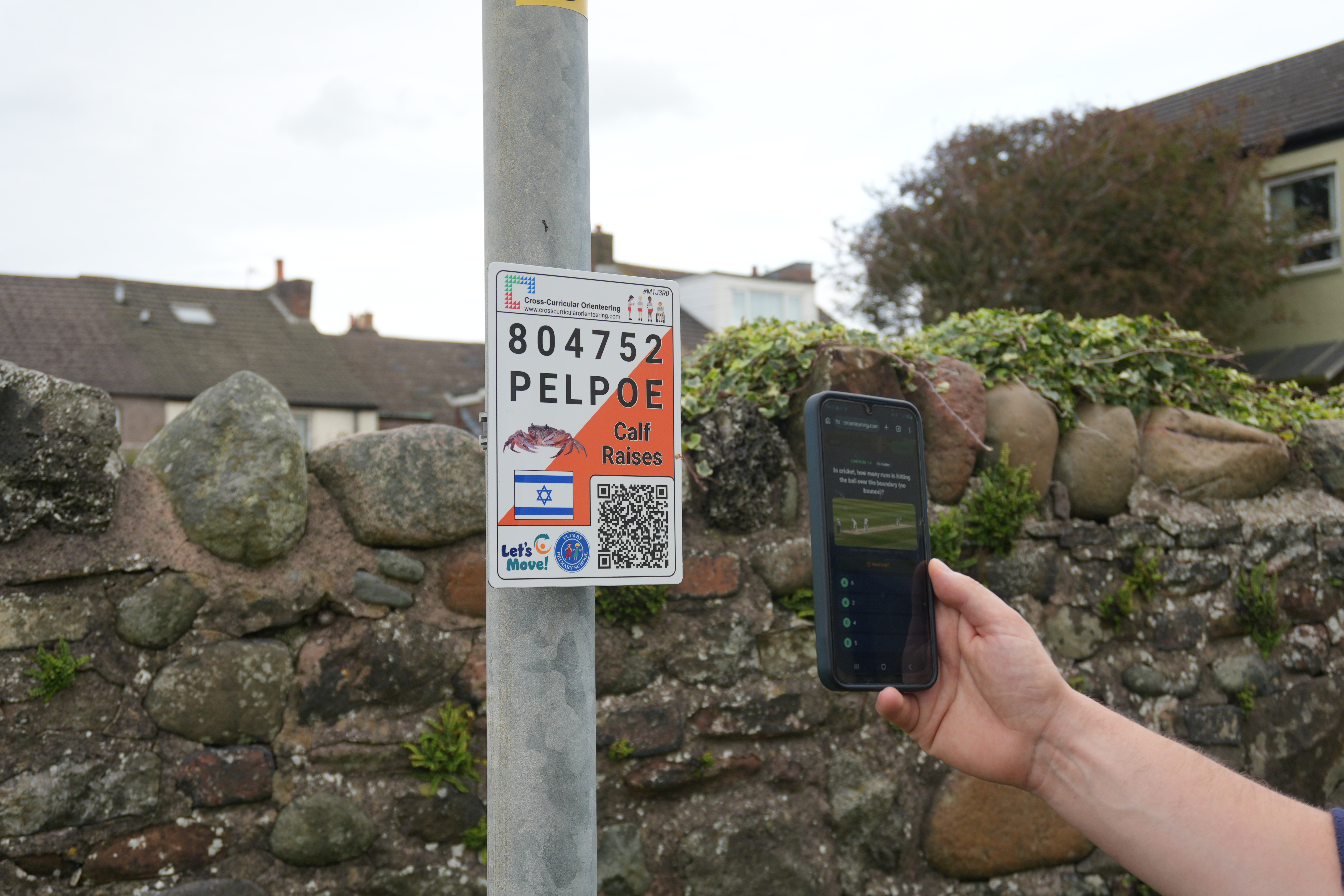 A person holds a smartphone beside an orienteering control marker attached to a lamppost. The marker features a unique code, QR code and educational clues, while the phone is being used to record a checkpoint on the community orienteering course. A stone wall, houses and greenery can be seen in the background.