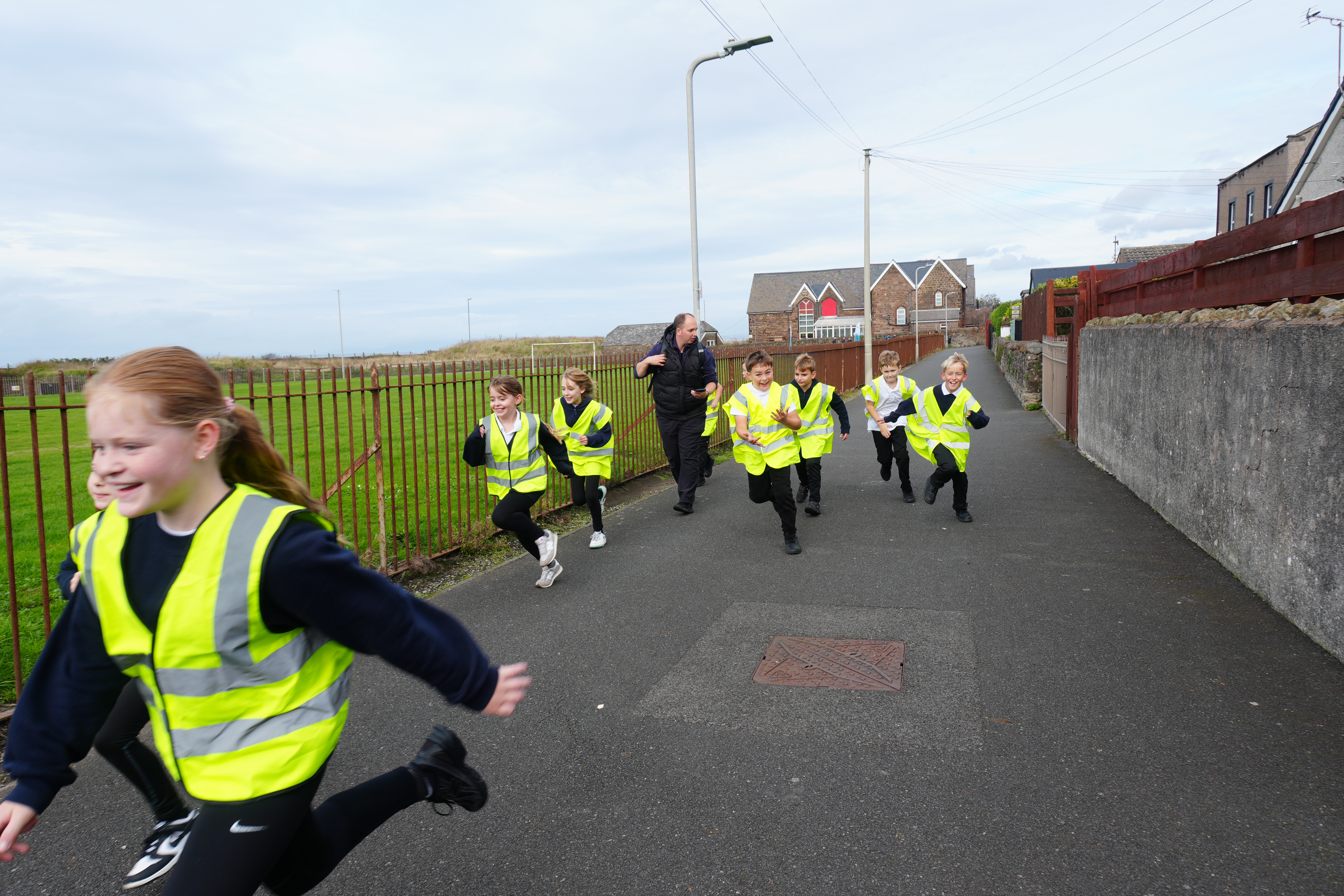 A group of primary school children wearing high-visibility vests run along a paved path during an outdoor orienteering activity. An adult walks with the group beside a fenced grass field, with houses and a residential street visible in the background. The activity takes place in a community setting under an overcast sky.