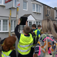 Children wearing high-visibility vests gather around an orienteering control marker attached to a street sign on a residential road, while accompanying adults observe. The group is taking part in an outdoor orienteering activity in a village setting with houses lining the street.