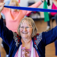 an older lady taking part in a seated exercise class. She is holding a blue exercise band above her head