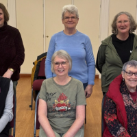 A group of 6 ladies from a chair based exercise class. 3 are standing, 3 are sitting.  They are smiling at the camera