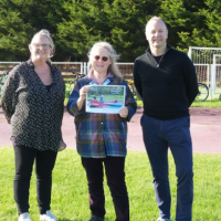 Three people stand on a grass field beside an outdoor cycle track. The person in the centre is holding a document. Several bicycles are visible in the background alongside a football goal and advertising boards, with trees lining the edge of the venue on a bright, sunny day.