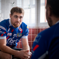 A rugby player sat in a changing room talking to another player who is sat opposite with his back to the camera