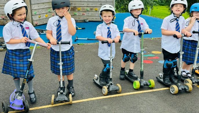 a group of children on scooters dressed  in school uniform wearing helmets in a row smiling the camera