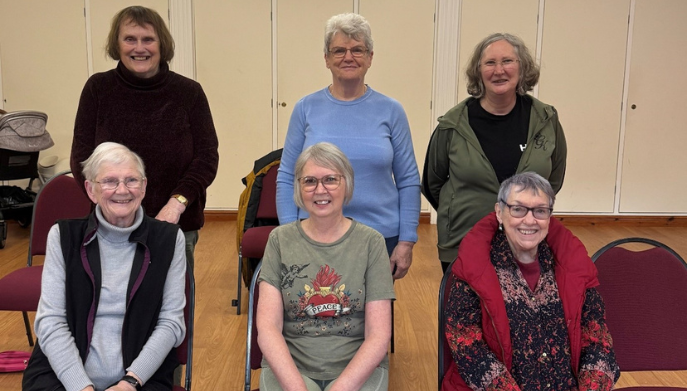 A group of 6 ladies from a chair based exercise class. 3 are standing, 3 are sitting.  They are smiling at the camera