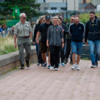 Group of adults walking together along a waterside path, with grassland on one side and buildings visible in the background.