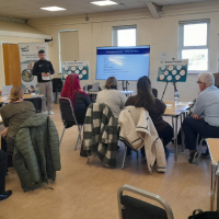 A meeting room with people sat at round tables.  There is a man stood at the front of the room presenting with a screen showing the text Telling the Story