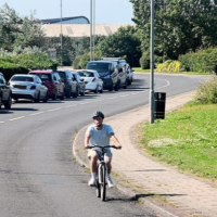 Person cycling along a roadside path beside parked cars and a grassy verge on a sunny day.