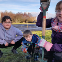 three children outdoors planting trees they are crouched down and smiling at the camera