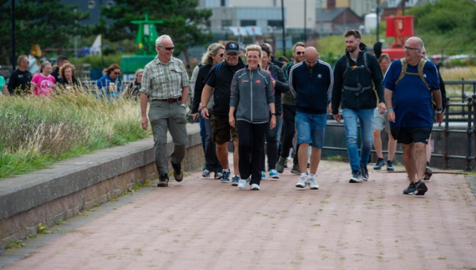 Group of adults walking together along a waterside path, with grassland on one side and buildings visible in the background.