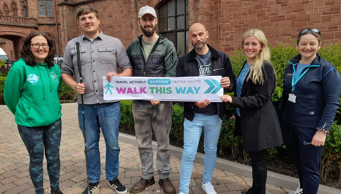 A group of adults stood smiling at the camera - they are holding a banner which reads: Travel Actively Barrow Invites You To Walk This Way