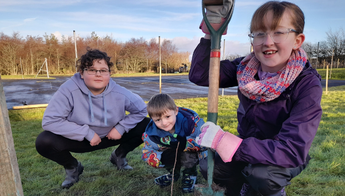 three children outdoors planting trees they are crouched down and smiling at the camera