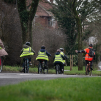a group of school children cycling on a road.They are wearing green hi-vis jackets.  They are following a cycle leader who is wearing an orange hi-vis jacket