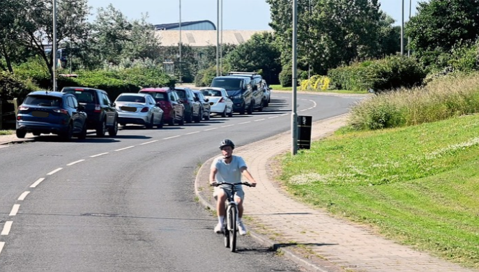 Person cycling along a roadside path beside parked cars and a grassy verge on a sunny day.