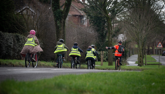 a group of school children cycling on a road.They are wearing green hi-vis jackets.  They are following a cycle leader who is wearing an orange hi-vis jacket