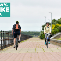 two people cycling along the channelside in Barrow.  White text on a green background reads Barrow's Big Bike Week